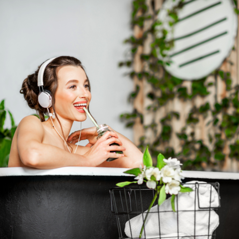 Junge entspannte Frau, die der Musik lauscht und Smoothie trinkt, während sie in der Retro-Badewanne im schönen Badezimmer mit grünen Pflanzen liegt